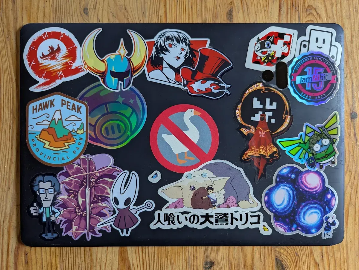 Black laptop cover covered in game and anime stickers, among them Hollow Knight, Persona 5, Untitled Goose Game, Journey and The Last Guardian