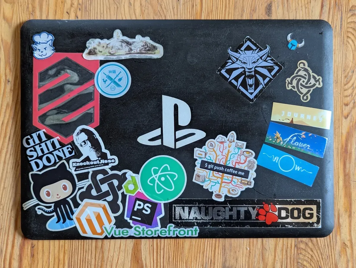 Worn black laptop cover with older stickers, among them PlayStation, Naughty Dog, GitHub, Magento, Vue Storefront, PhpStorm and Atom